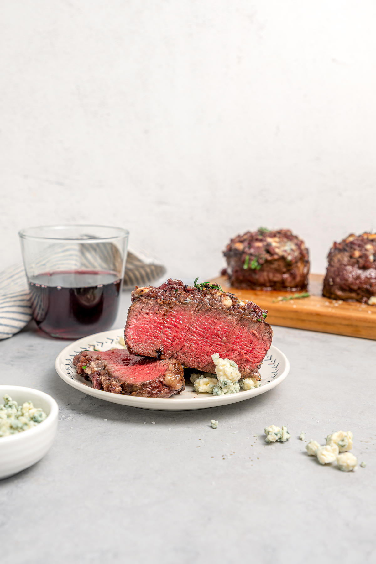 A glass of red wine next to a wooden board with blue cheese crusted steak. One piece of steak has been served on a small plate.