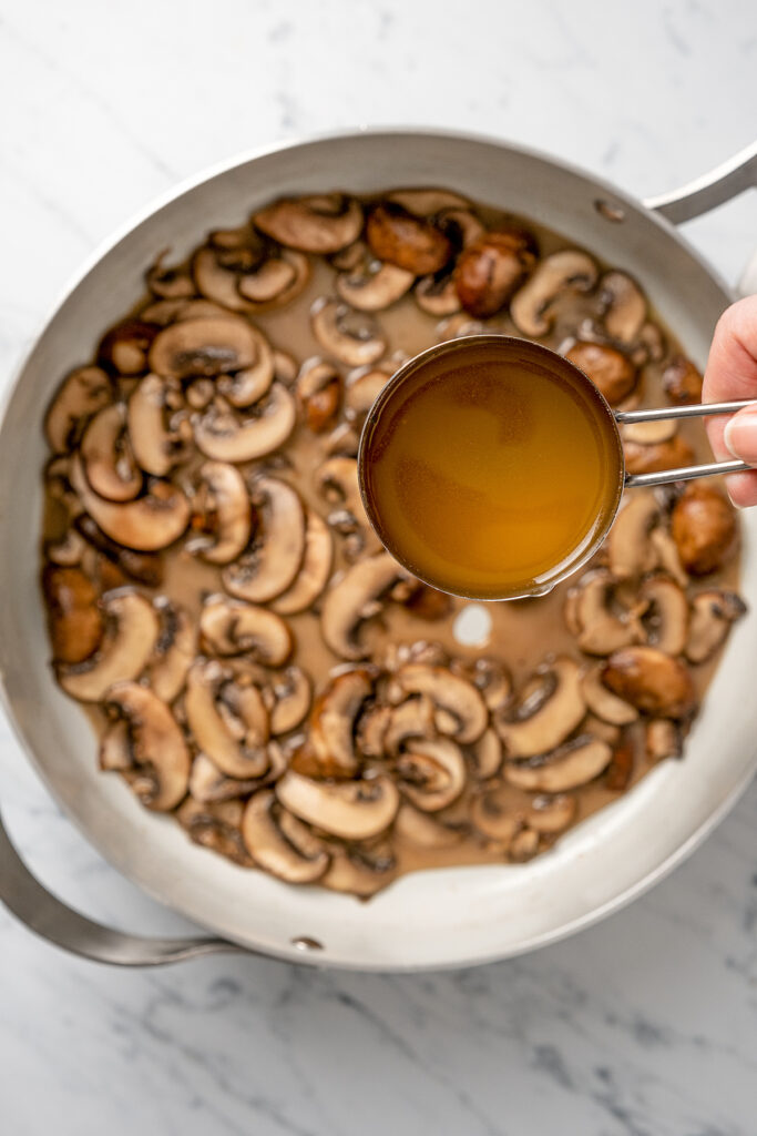 Chicken broth being poured into a pan of sauteed mushrooms.