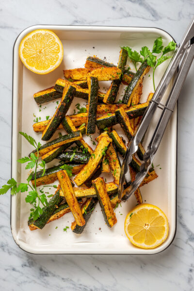 Baked zucchini fries on a large tray with lemon halves.