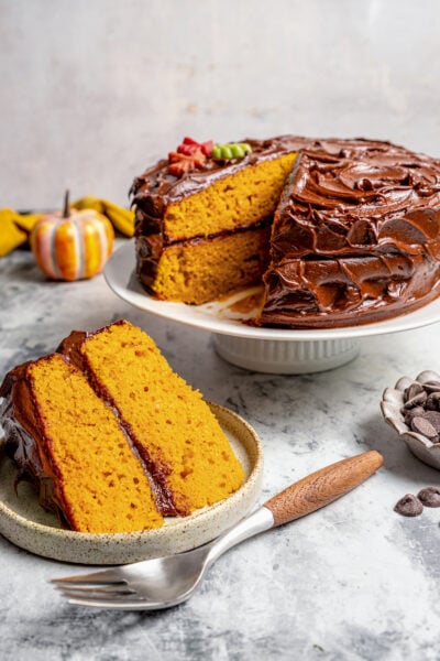 A slice of pumpkin cake with chocolate frosting on a serving plate, and the rest of the cake in the back.