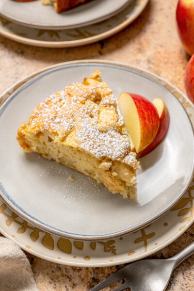 A slice of fluffy apple cake with powdered sugar on top and sliced apples on the side.