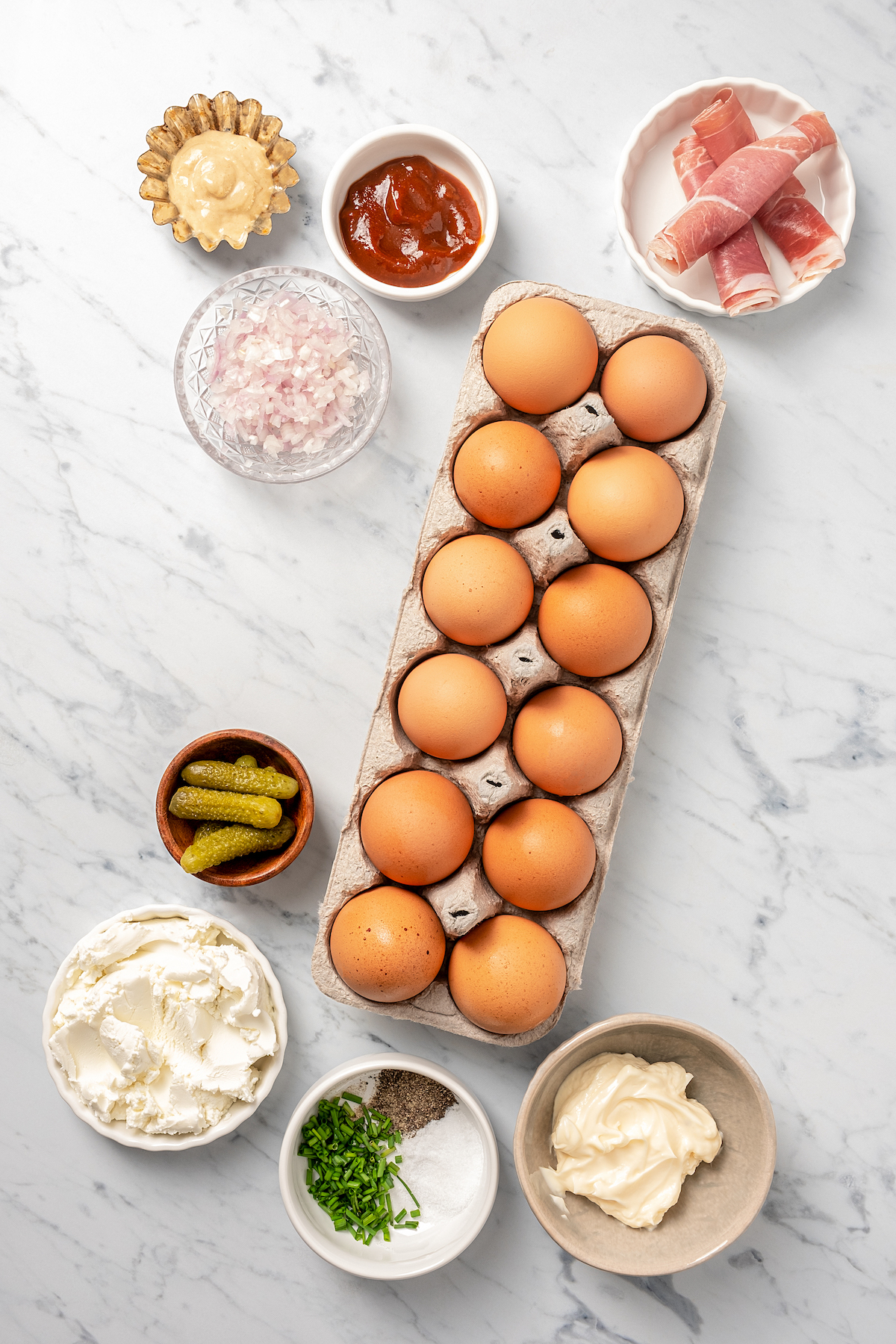 Eggs, sriracha, prosciutto, cornichons, seasonings, mayonnaise, and other ingredients on a work surface.