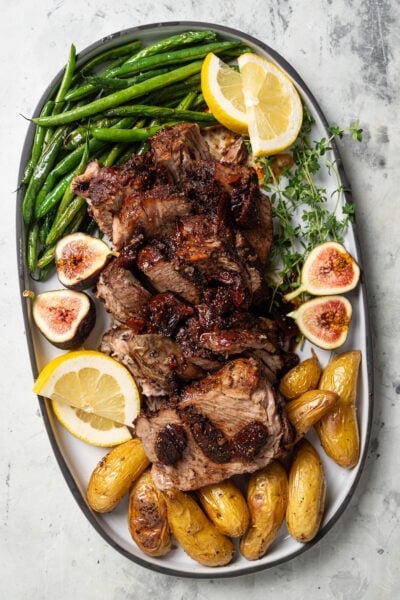 Sliced pork shoulder recipe on a serving plate with green beans and potatoes on the side.