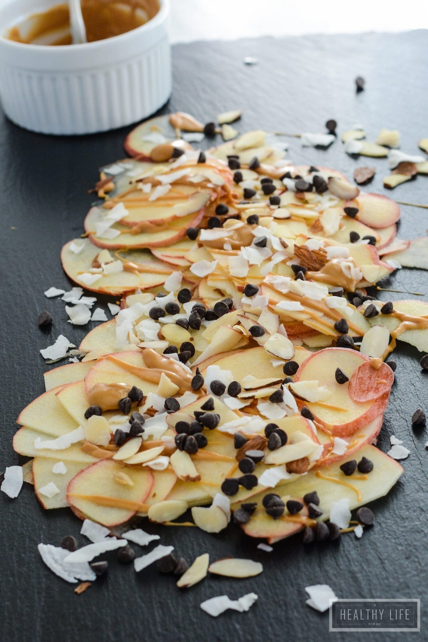 Thinly sliced apples laid out on a table with almond butter, sliced almonds, flaked coconut and mini chocolate chips on top