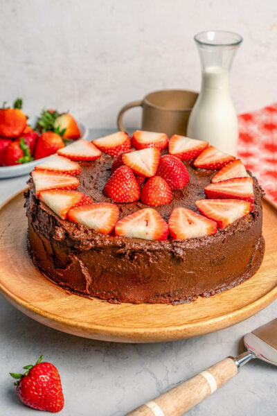 Angled photo of a whole cake with chocolate frosting and sliced strawberries.