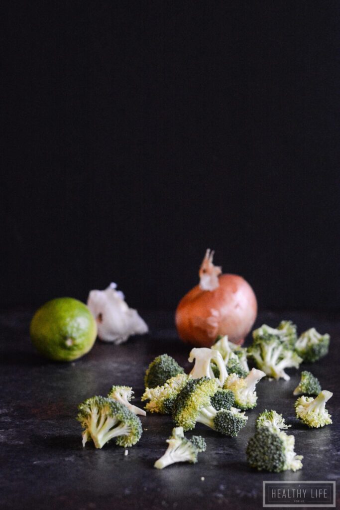 A black countertop with broccoli florets, an onion, garlic, and a lime.