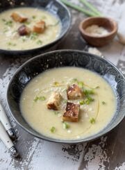 A close up of a bowl of potato leek soup garnished with croutons, olive oil, parmesan cheese, and chives