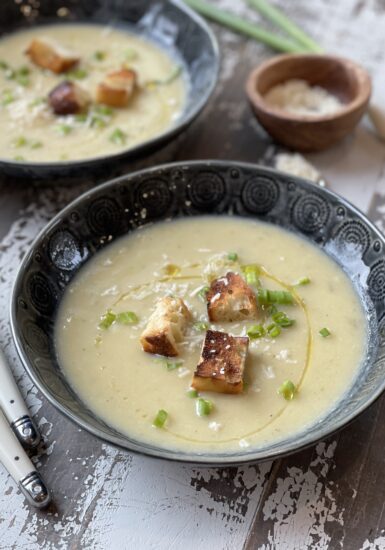 A close up of a bowl of potato leek soup garnished with croutons, olive oil, parmesan cheese, and chives