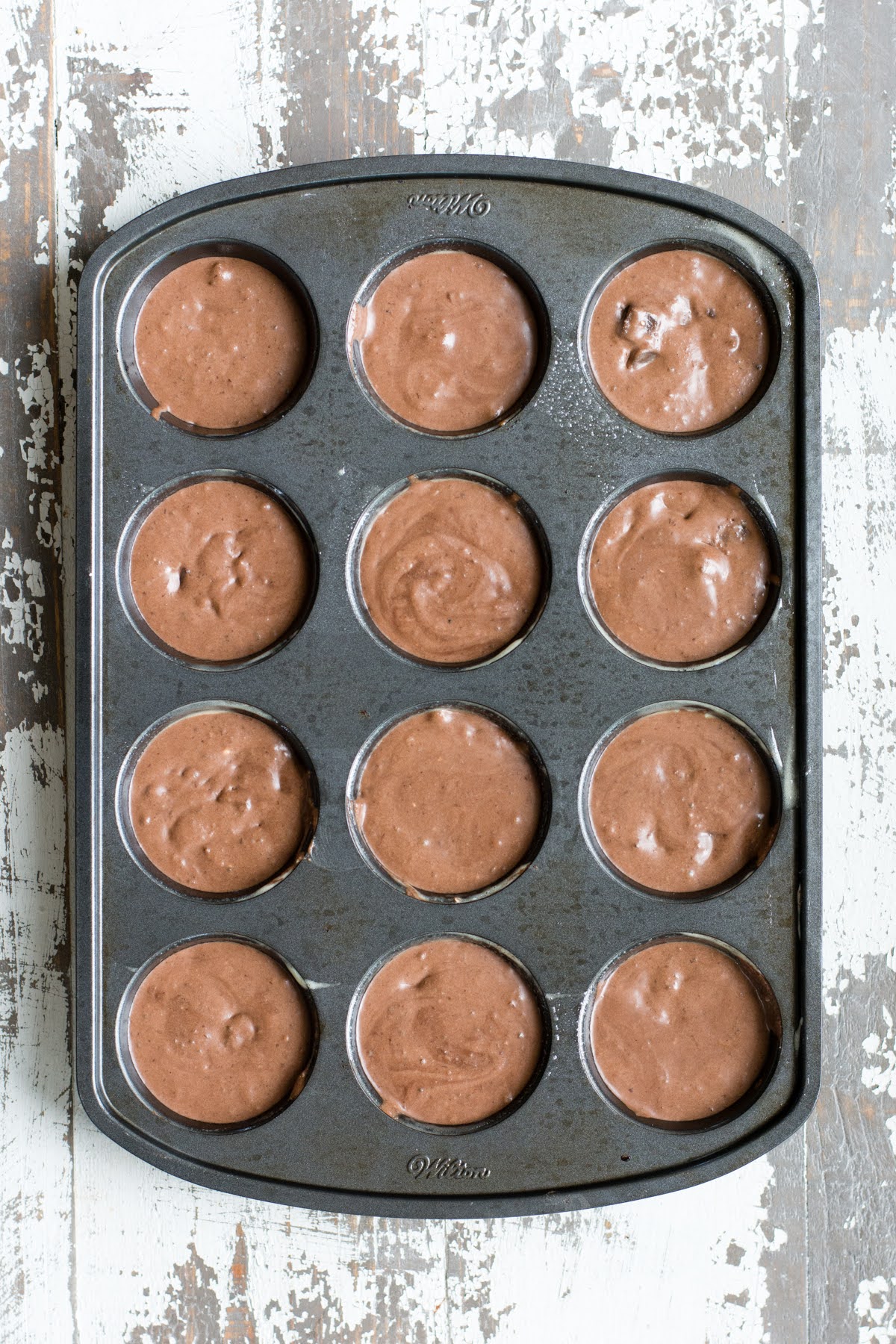 A cupcake pan on a dining table with all 12 liners filled with muffin batter