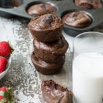 A stack of 3 double chocolate protein muffins beside another half muffin and a glass of plant-based milk