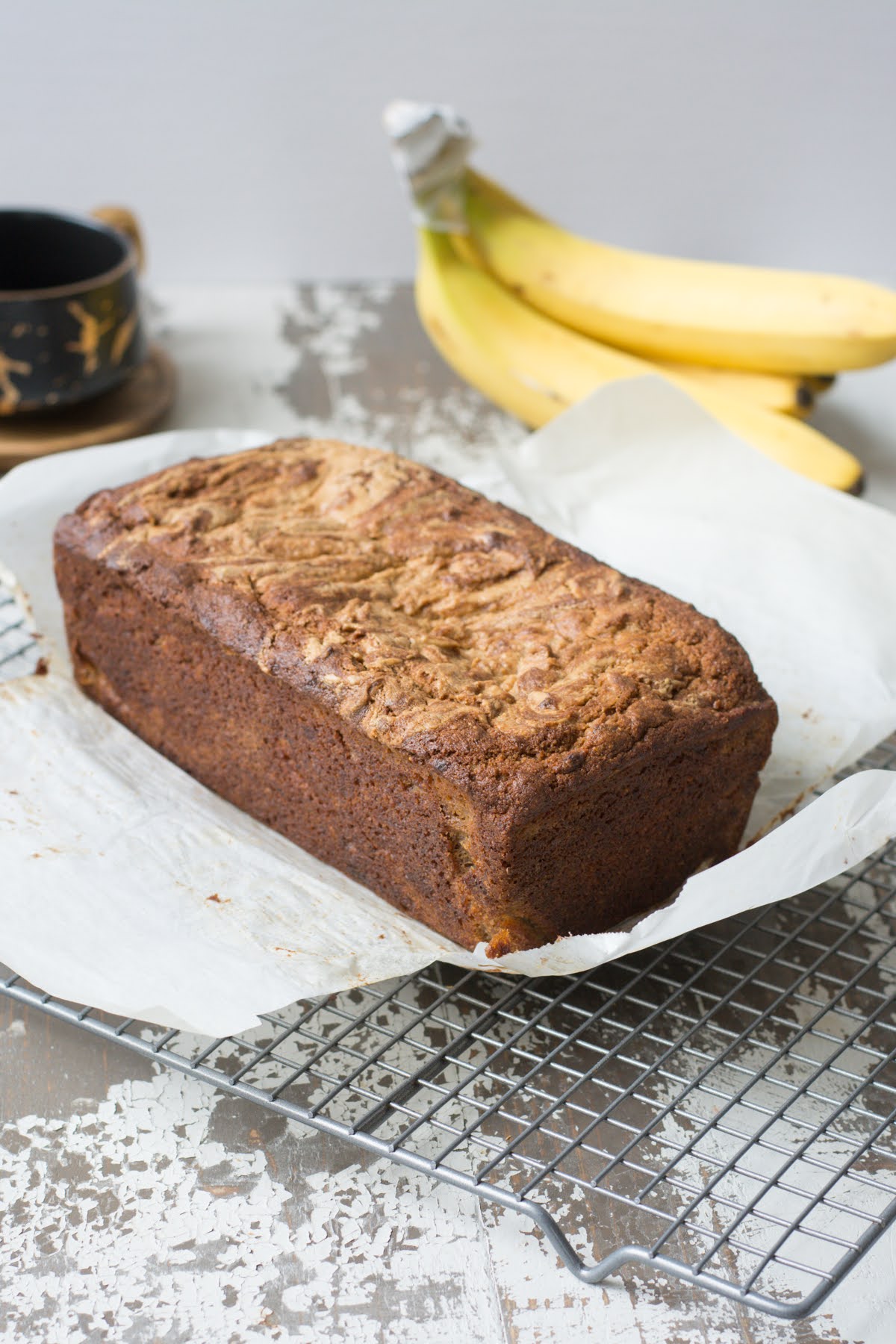 A loaf of banana peanut butter bread on a wire rack with fresh bananas in the background