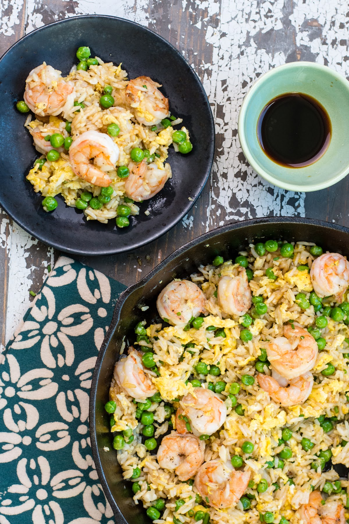 A skillet full of shrimp fried rice beside a single serving in a shallow bowl and a small dish of soy sauce