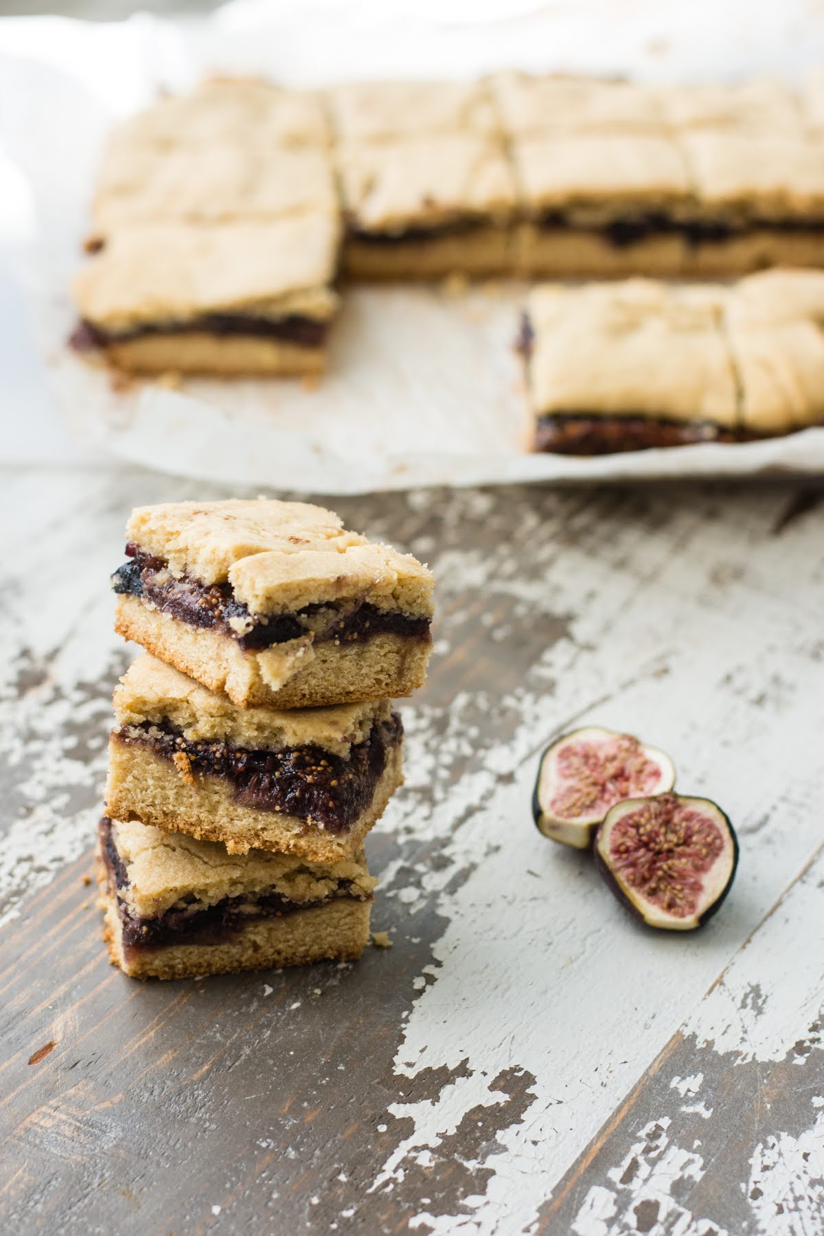 Homemade fig bars stacked up in front of the rest of the batch. A halved fig lies nearby.
