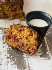 A slice of cranberry loaf cake next to a cup of milk. The remainder of the loaf is on the table in the background.