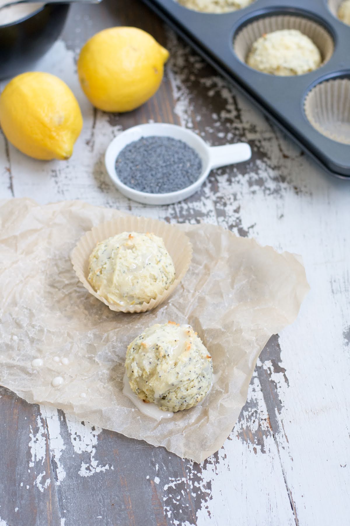 Two small muffins on a piece of parchment, next to a dish of poppy and two lemons.