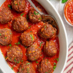 Overhead shot of a white baking dish of meatballs with a striped cloth napkin, herbs, and sauce.