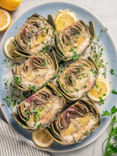 Halved artichokes with garlic, fresh herbs, and lemon wedges.